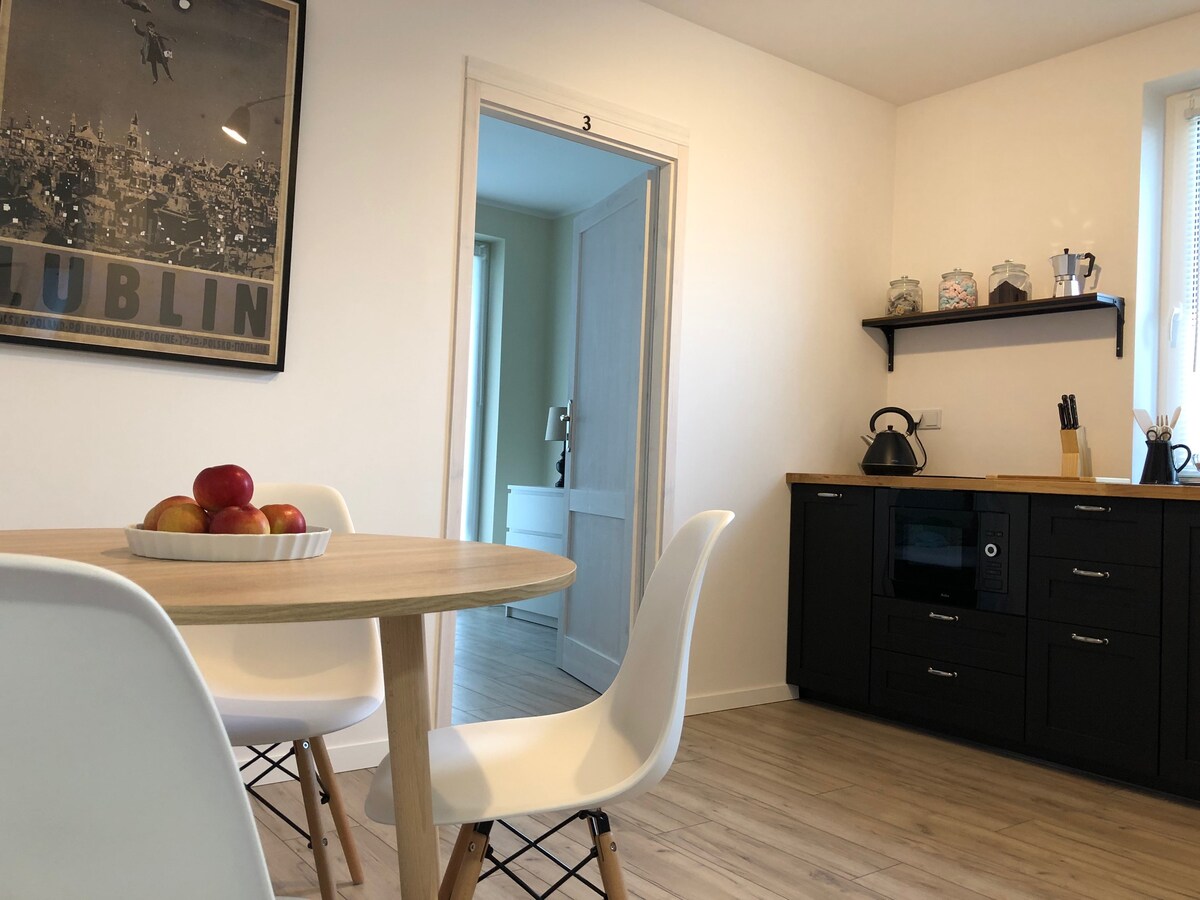 A dining area features a round wooden table set with a bowl of apples. White chairs surround the table, while a kitchenette with dark cabinetry and modern appliances is visible in the background. A poster of Lublin adorns the wall, adding a local touch.