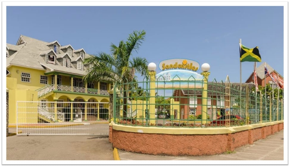 The exterior of SandCastle Resort is displayed with a vibrant yellow facade and decorative railings. A welcoming entrance is framed by lush greenery and palm trees. The Jamaican flag and other national flags are prominently displayed, providing a sense of place.