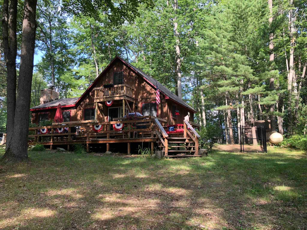 A chalet-style home is set among tall trees, with a spacious wooden deck adorned with decorative elements. The entrance is visible with steps leading up to the main floor and a glimpse of a fenced yard on one side. Natural greenery surrounds the property.