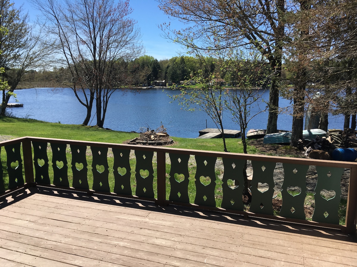 A deck overlooks a serene lake, framed by a patterned railing featuring hearts. Lush green grass leads to the water's edge, where trees provide shade. The surface of the lake reflects the clear blue sky, creating a peaceful and inviting outdoor space.