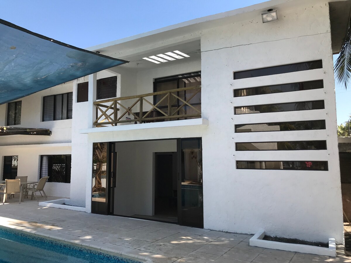 The exterior of the house features a two-story white structure with large glass doors providing access to the pool area. A balcony is visible on the upper level, and the design includes horizontal slats allowing for natural light.
