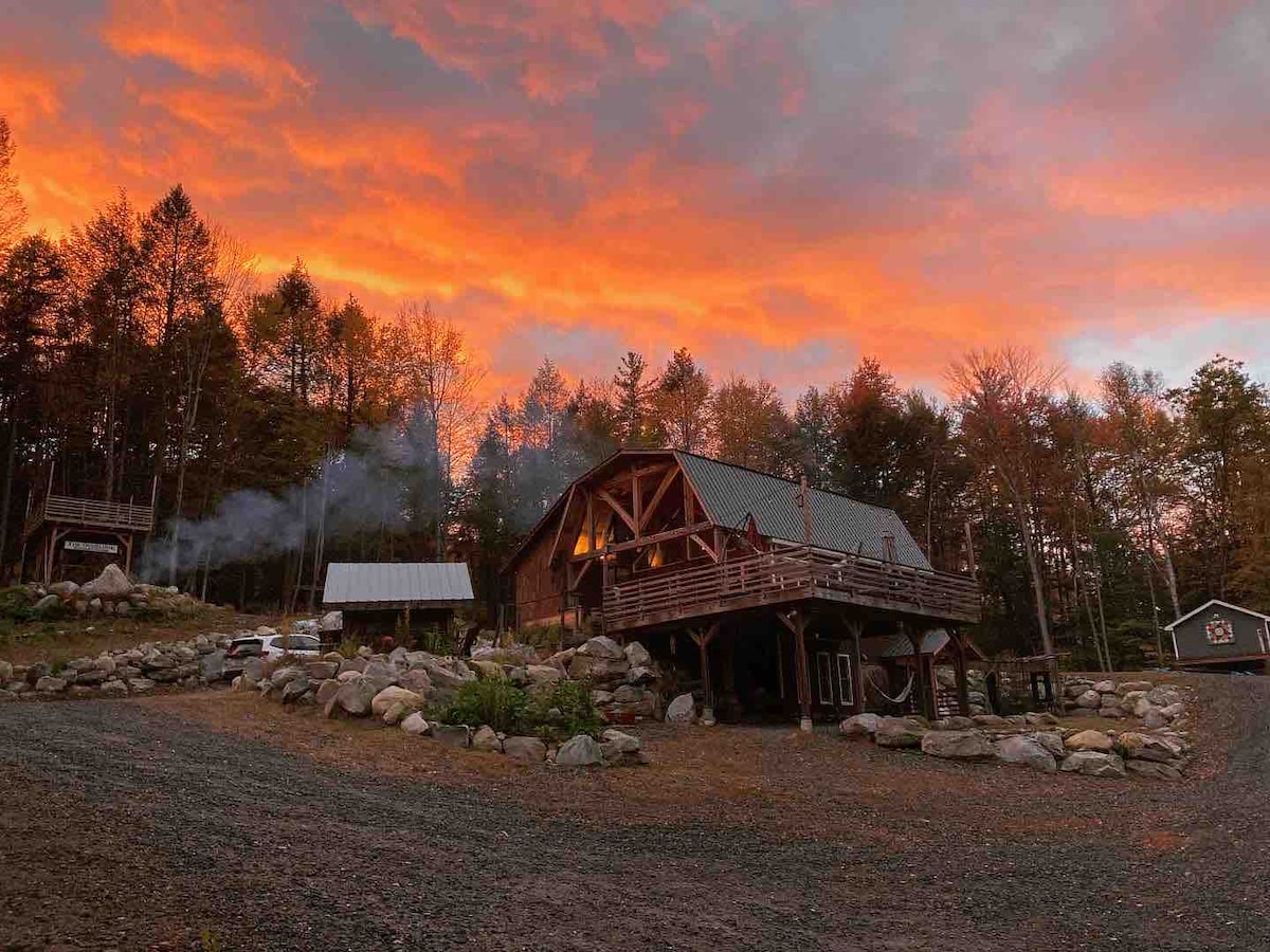 The house is set against a vibrant sunset sky, showcasing rustic architecture with wooden beams and multiple decks. Smoke rises gently from the chimney, while a natural stone landscape surrounds the property, enhancing the serene outdoor environment.