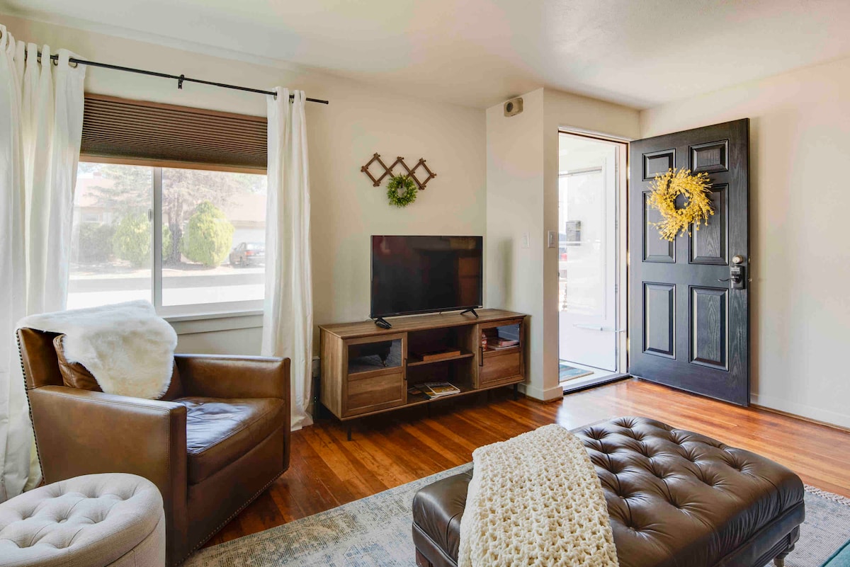A welcoming living area features hardwood flooring, a cozy armchair, and a modest entertainment unit holding a television. Natural light filters through the large front window, complemented by sheer white curtains. The front door is adorned with a decorative wreath, enhancing the inviting atmosphere.