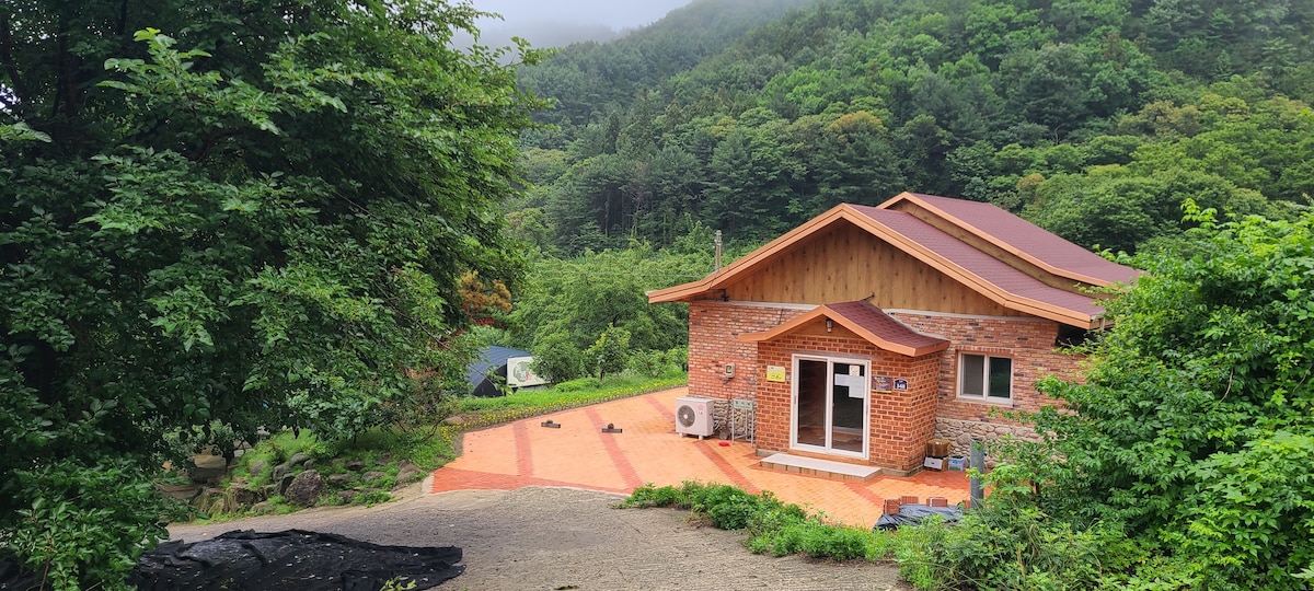 A brick house with a wooden roof is set against a backdrop of lush green mountains. A spacious outdoor area with a patterned floor is visible in front of the house. Surrounding trees provide natural shade, contributing to the tranquil setting.