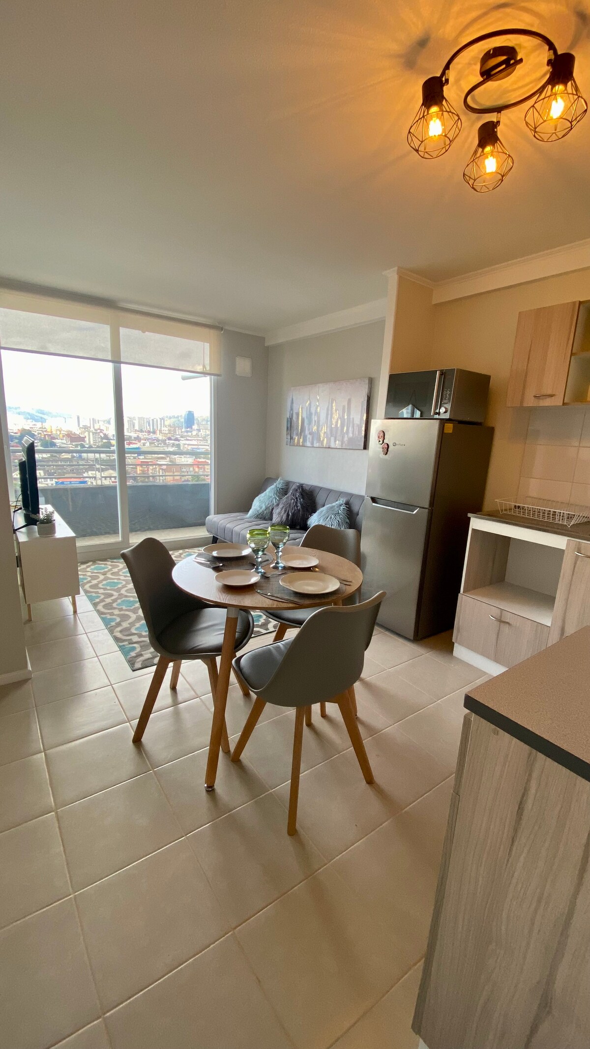 A modern living space features a dining table set for four alongside a comfortable gray sofa. A large window offers city views, while a small balcony is visible. Kitchen appliances, including a refrigerator and cabinets, are neatly arranged in the background.