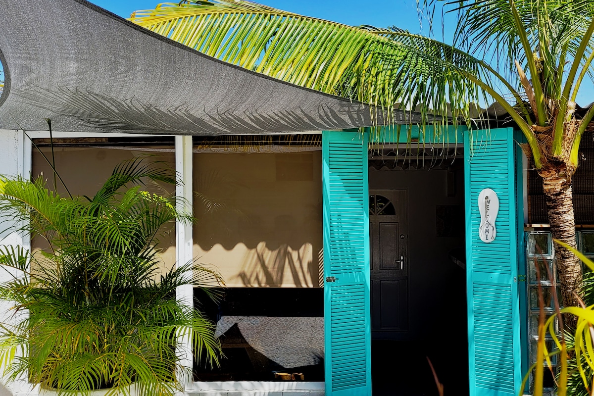 The entrance of a guesthouse is framed by vibrant turquoise shutters and a protective awning. Tropical plants are positioned nearby, creating a relaxed atmosphere. A welcoming door leads into the living space, which is illuminated by natural light.