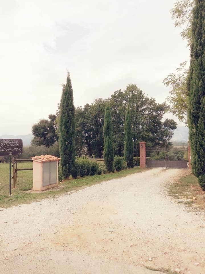 A gravel driveway leads towards a gated entrance, flanked by tall cypress trees and greenery. A sign indicating the residence is visible near the entrance, surrounded by a peaceful landscape that hints at the natural beauty of the Tuscan countryside.