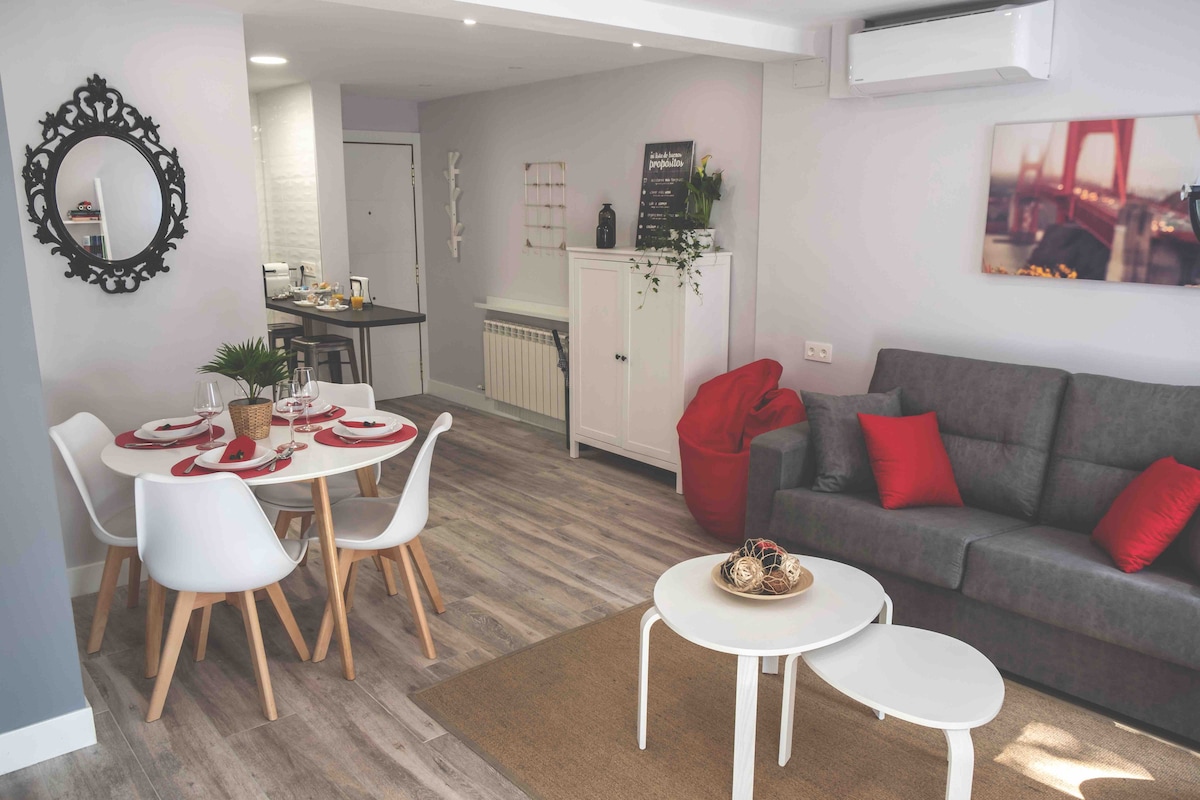 A modern living space is showcased, featuring a gray sofa with red accents and a glass coffee table. A dining area with white chairs surrounds a circular table set for four. Light wood flooring complements the neutral wall tones, enhancing the room's spacious feel.
