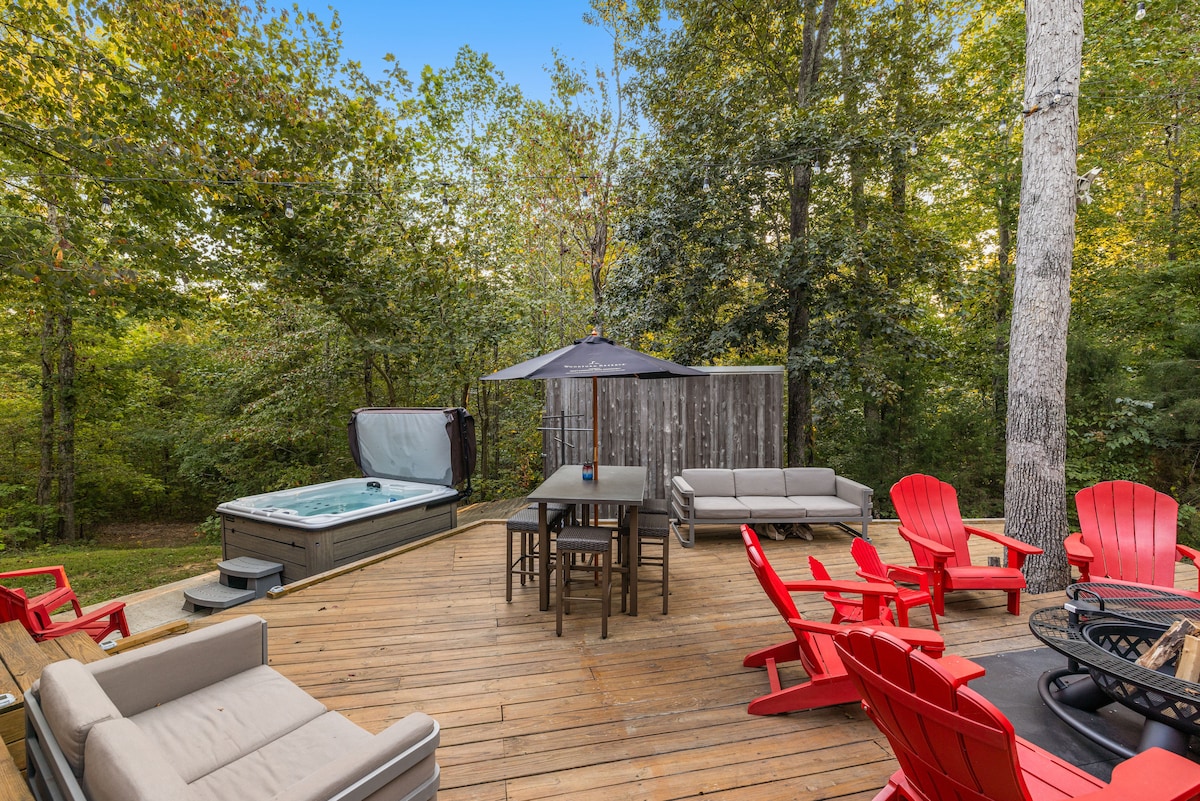 A spacious wooden deck surrounded by trees features a hot tub and comfortable seating arrangements. Red adirondack chairs and a large dining table create inviting spaces for relaxation and entertaining. An umbrella provides shade for outdoor gatherings.