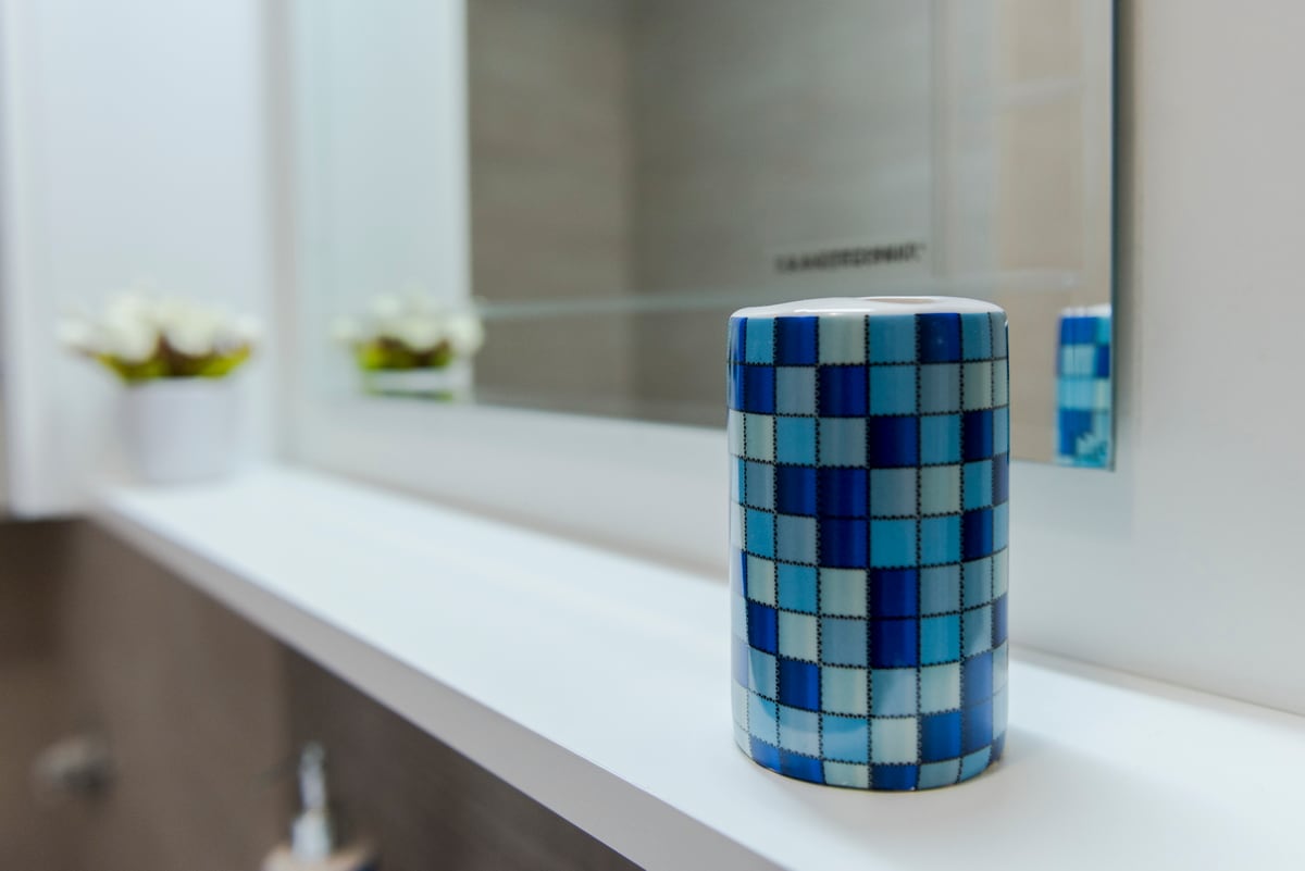 A vibrant blue mosaic bathroom cup is positioned on a white shelf, reflecting light from the nearby mirror. Soft white flowers in a small pot are placed in the background, adding a touch of nature to the modern bathroom setting.