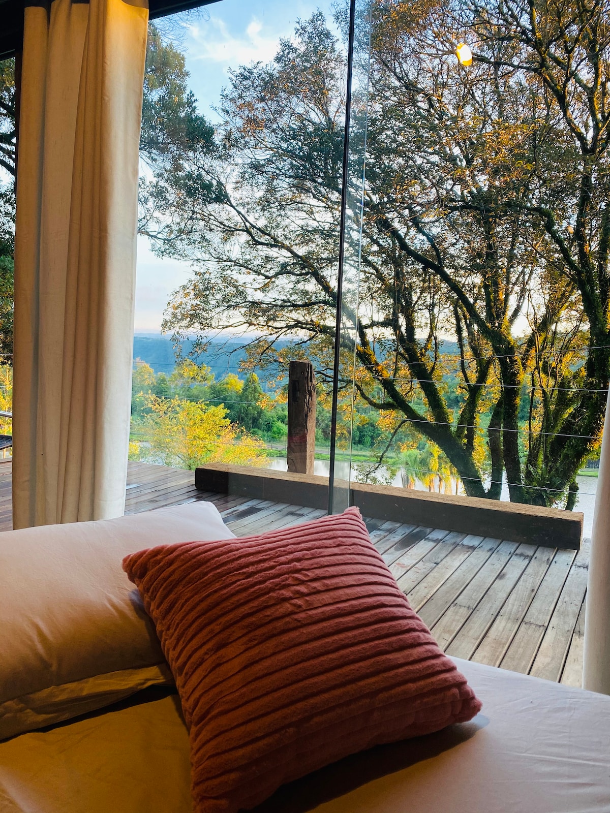 A spacious bed is adorned with a textured pink pillow, positioned by large glass windows. Through the windows, vibrant trees and rolling hills can be seen, reflecting the changing colors of the season under natural light.