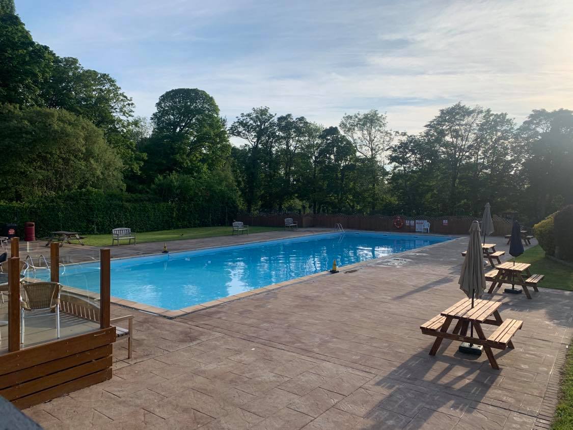 An outdoor swimming pool is surrounded by a spacious deck featuring wooden picnic tables and umbrellas. Lush trees provide shade around the area, enhancing the pool's inviting blue water, ideal for relaxation during warmer months.