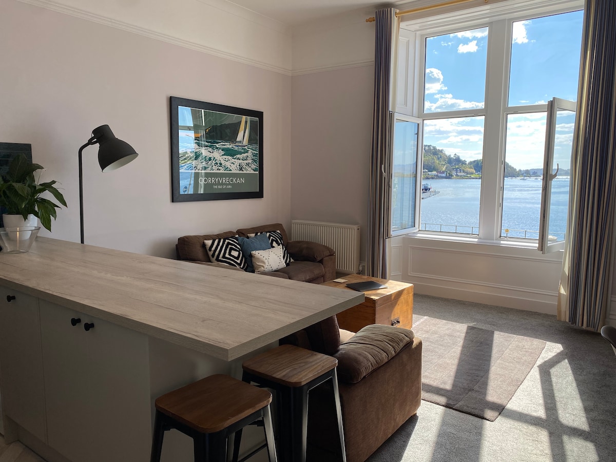 The open-plan living area features large windows that provide sweeping views of Oban Bay. A cozy seating arrangement includes a spacious settee, complemented by a stylish coffee table. A dining island with stools encourages casual dining while enjoying the scenery.