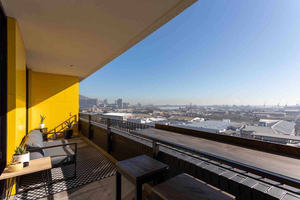 A spacious balcony is featured with outdoor seating and small tables. Bright yellow walls create a vibrant setting, while sweeping views of the city and ocean are visible under a clear blue sky. The area is perfect for enjoying drinks or socializing.