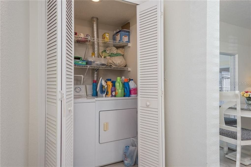 A utility closet is depicted, featuring a stacked washer and dryer. Shelves hold a variety of cleaning supplies, laundry detergents, and other household items, contributing to a functional laundry area.
