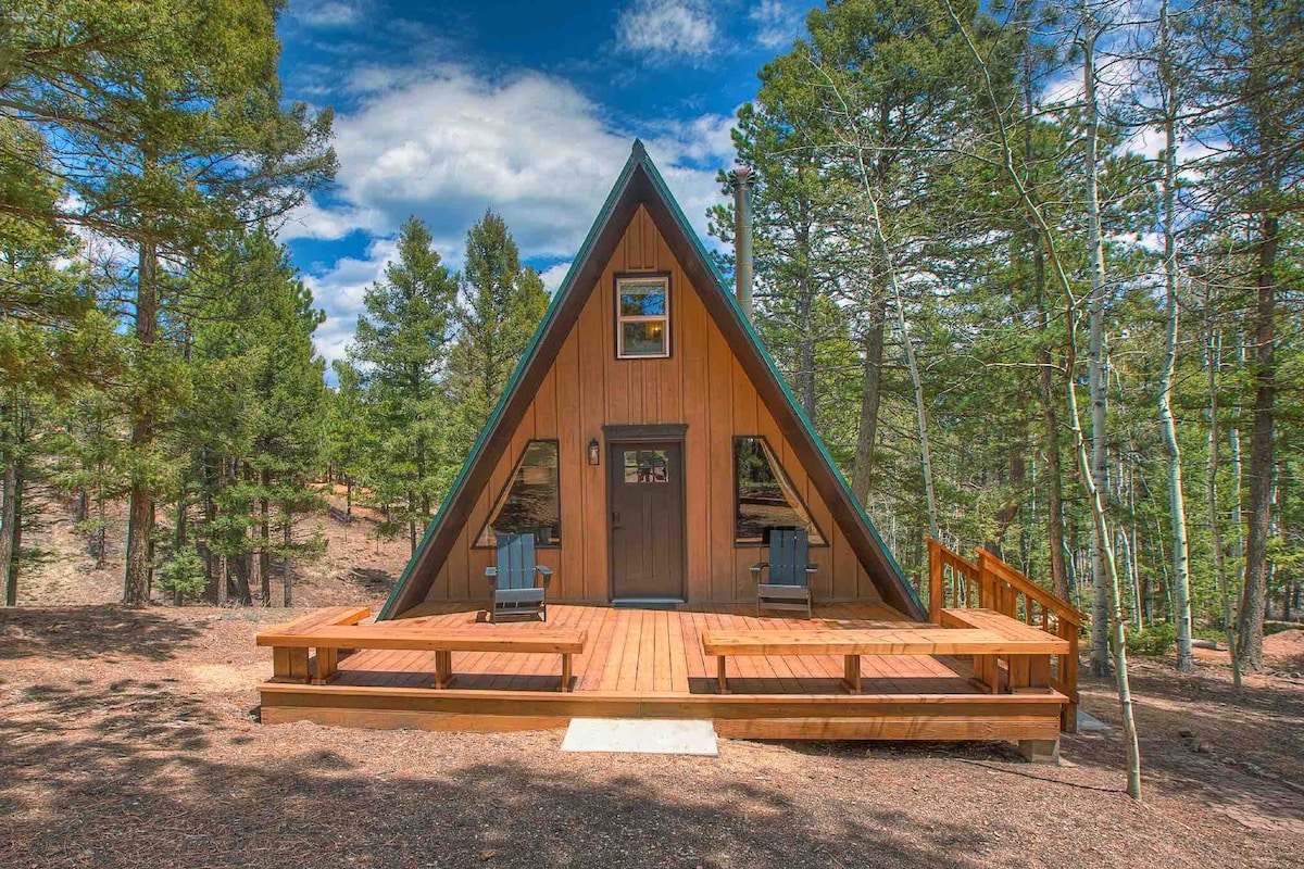 The exterior of the A-frame cabin is surrounded by tall trees, featuring a wooden deck with two adirondack chairs. A large triangular window adorns the front, complemented by a welcoming entrance door, emphasizing the cabin's charming architecture.