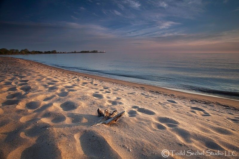 A serene beach landscape is captured at dawn, featuring smooth sandy shores that gently curve towards a calm lake. Footprints are visible in the soft sand, leading towards the water's edge, while a few scattered driftwood pieces are placed along the shoreline.