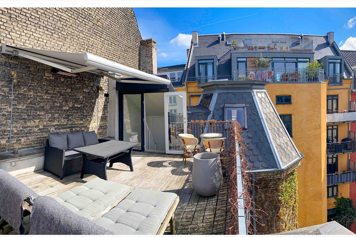 A private roof terrace is shown, featuring comfortable lounge seating and a table. The wooden deck is surrounded by a low wall, providing views of neighboring rooftops and greenery. Bright sunlight illuminates the space, enhancing its inviting atmosphere.