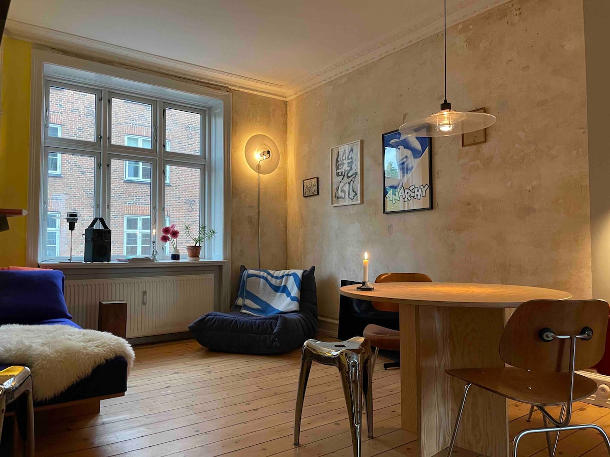Natural light fills a cozy living space featuring rustic walls and a well-lit dining area. A round table with wooden chairs is positioned beside a comfortable lounge chair. Vibrant artworks adorn the walls, enhancing the room's artistic charm.
