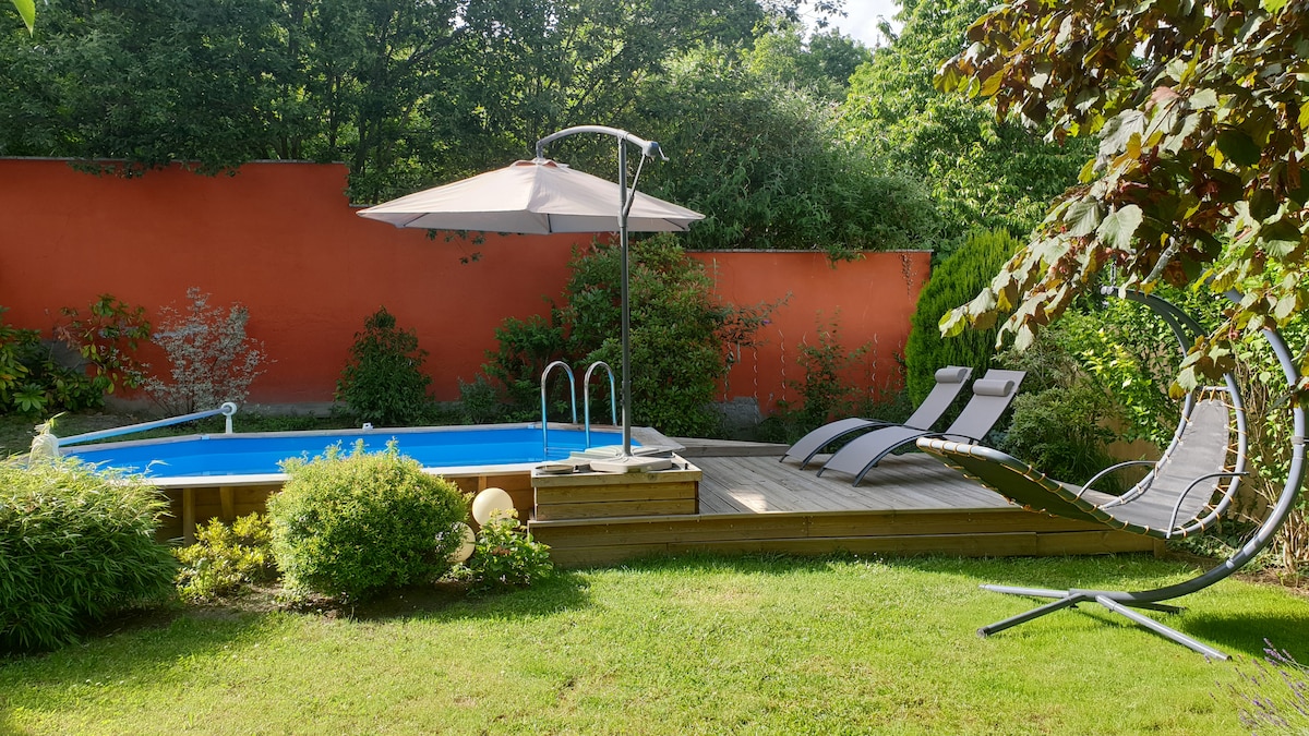 A private outdoor area features a swimming pool surrounded by wooden decking. Two lounge chairs are positioned for relaxation under a large umbrella. Lush greenery and a vibrant red wall provide a serene backdrop, with a gently curved swing chair adding to the inviting space.