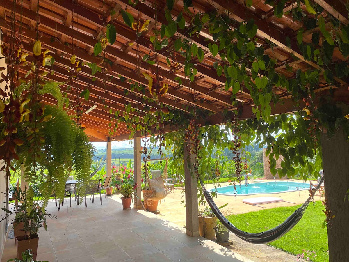 A covered patio features a wooden ceiling and is adorned with hanging plants. A hammock is suspended nearby, and a swimming pool is visible in the background, surrounded by grassy areas and flower arrangements. Tables and chairs are set for outdoor enjoyment.