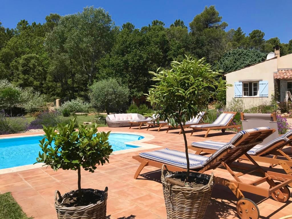 A serene outdoor space features a sparkling swimming pool surrounded by sun loungers with striped cushions. Potted trees add greenery, while the charming gite is partially visible in the background, complemented by lush trees and blue skies.