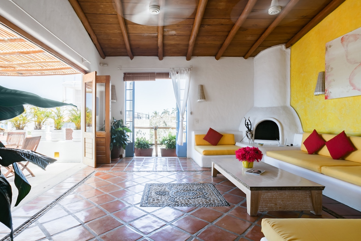 An inviting living area features a combination of yellow walls and rustic wooden beams. Comfortable seating is arranged around a central coffee table adorned with vibrant flowers. Sunlight illuminates the space, highlighting the terracotta tiled floor and the large windows that offer a view of the outdoors.