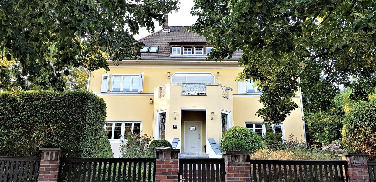 The villa, with a classic yellow facade, features distinct architectural details including a balcony and multiple windows framed by greenery. A welcoming entrance is highlighted by a set of stairs leading to the front door, bordered by well-maintained gardens.