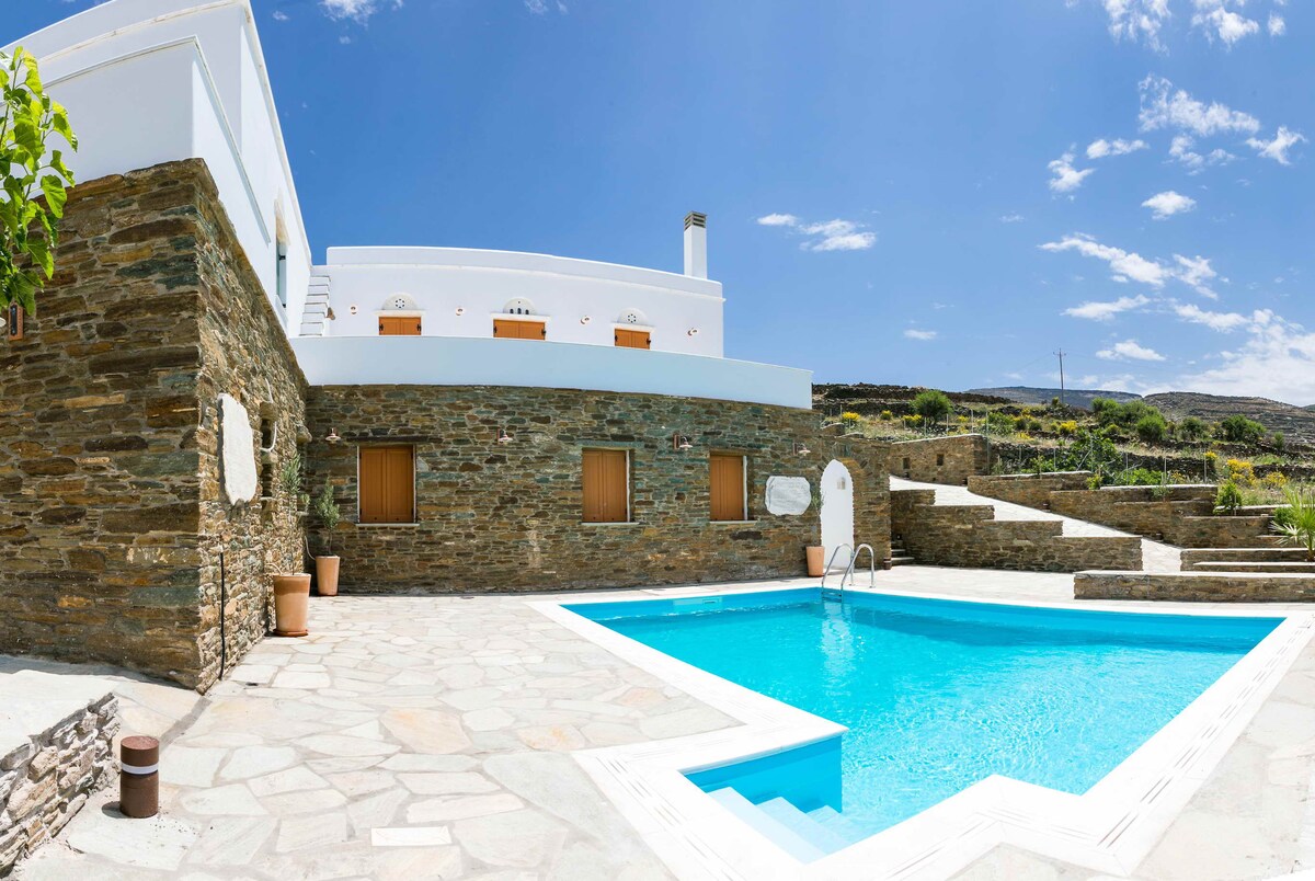 The outdoor space features a modern swimming pool surrounded by a stone patio. The villa's exterior is characterized by clean lines and neutral tones, with several windows allowing natural light. Lush greenery complements the serene atmosphere, and the sky is clear blue, enhancing the environment.
