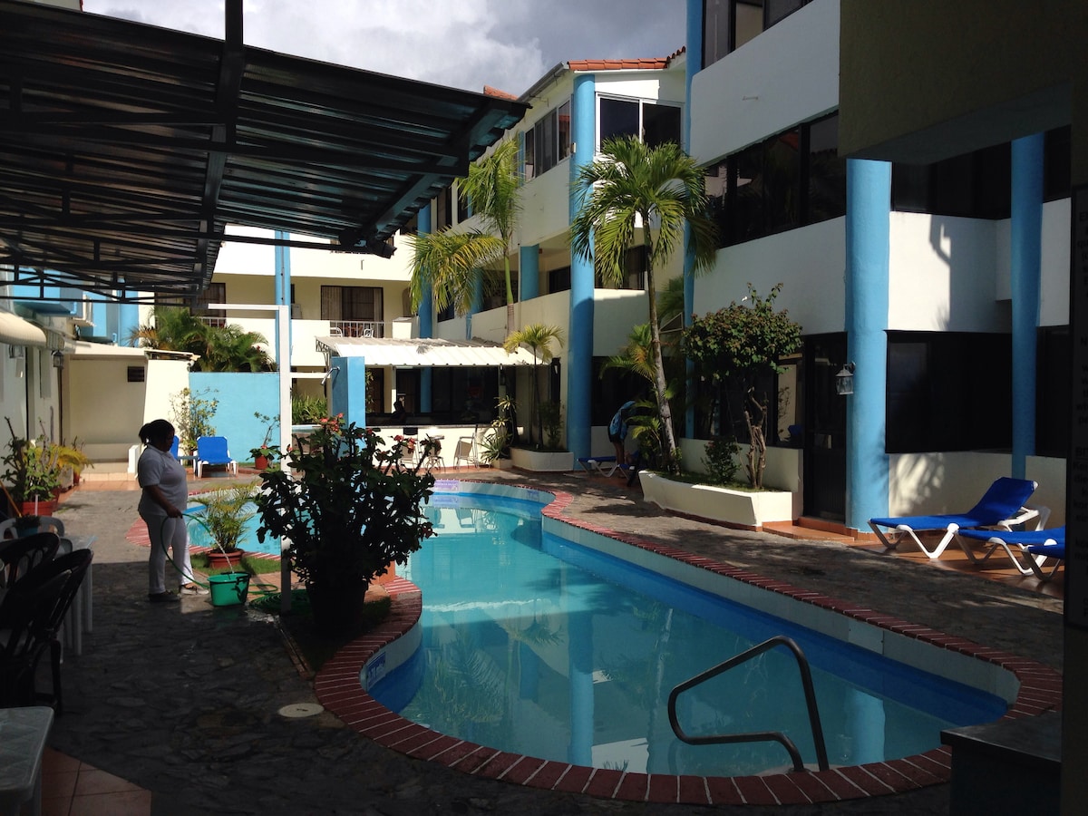 The swimming pool is framed by a tropical courtyard, featuring palm trees and lush greenery. Lounge chairs are positioned around the pool area, which is partially shaded by awnings. The surrounding blue and white building creates a bright and inviting atmosphere.