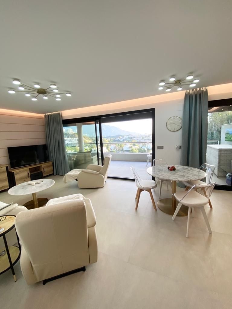 A modern living area showcases large glass doors that open to a scenic balcony. Comfortable cream-colored armchairs are paired with a circular coffee table. A marble dining table with transparent chairs complements the space. Soft lighting creates a serene atmosphere, while a clock adorns the wall.