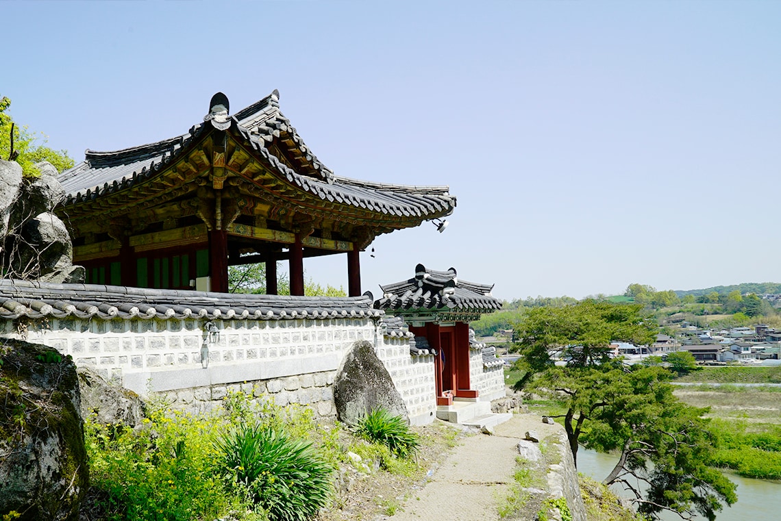 A traditional building with a curved, tiled roof and wooden accents is situated on an elevated area. The structure is surrounded by natural greenery and large stones, with a scenic view of the river and village in the background. Clear skies frame the scene.