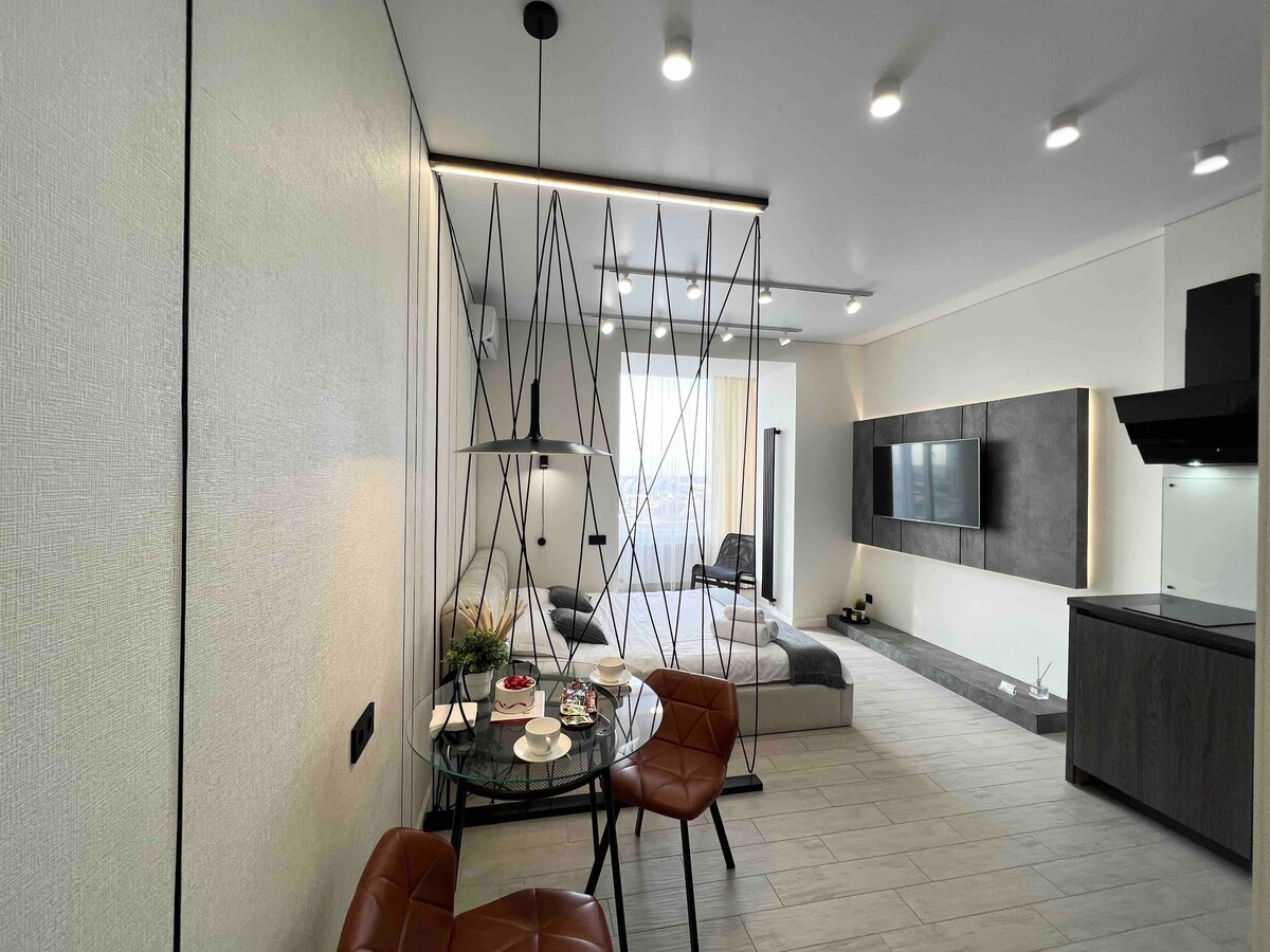 A modern studio features a dining area with a round table set for two, surrounded by stylish chairs. Soft lighting highlights the open space, which includes a comfortable seating area and large windows that let in natural light. A sleek kitchen is visible in the background.