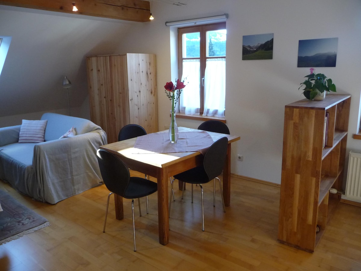 A light-filled living space features a wooden dining table surrounded by four black chairs. A comfortable sofa is draped with a light cover. Natural wooden cabinetry is displayed along with a flowering plant on the shelf. Windows allow views of the outdoors.
