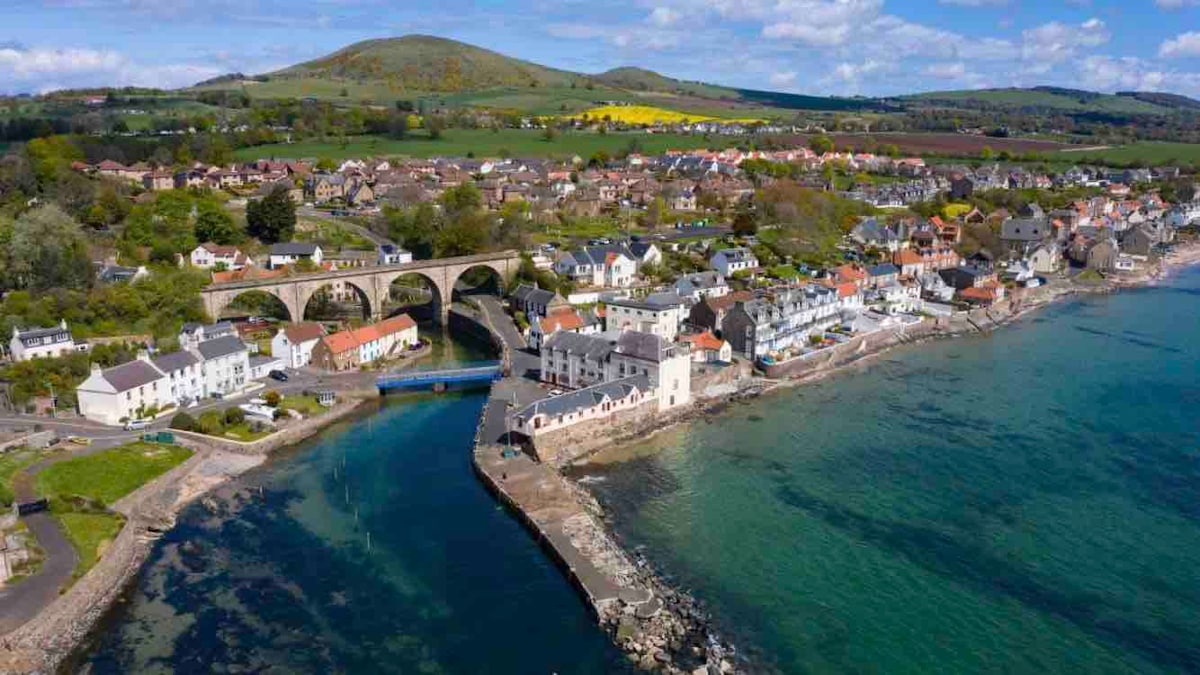 An aerial view of Lower Largo showcases a charming coastal village. Colorful houses line the seafront, with a notable stone bridge in the background. The clear waters gently lap against the shoreline, while rolling green hills provide a serene backdrop to the picturesque landscape.
