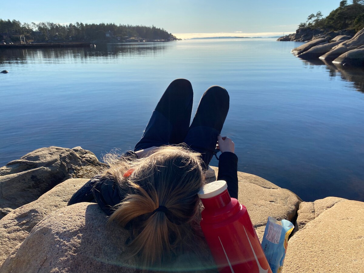 A person lounges on a large rock by the calm sea, with clear blue water stretching into the distance. A bright red water bottle rests beside them, contributing to a peaceful outdoor scene framed by trees in the background.