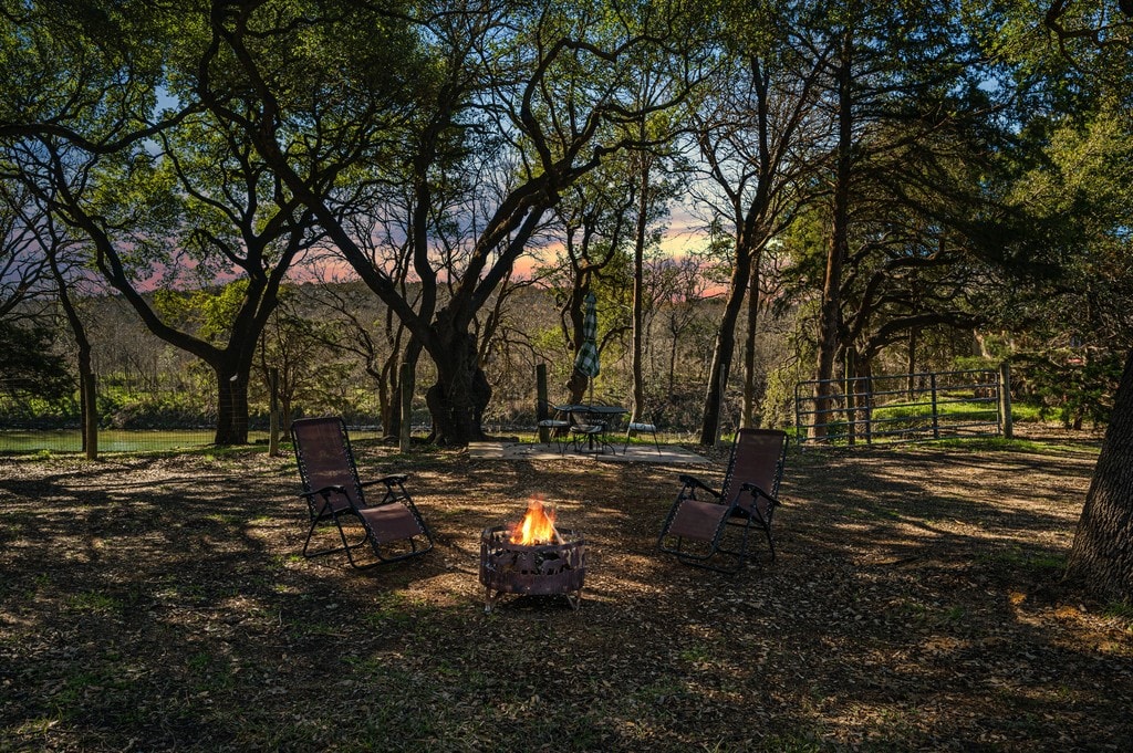 A tranquil fire pit area is surrounded by tall trees, offering a natural setting for gathering. Two lounge chairs are positioned comfortably by the fire, which glows warmly, inviting cozy evenings. The backdrop features a view of the lush landscape and a colorful sunset sky.
