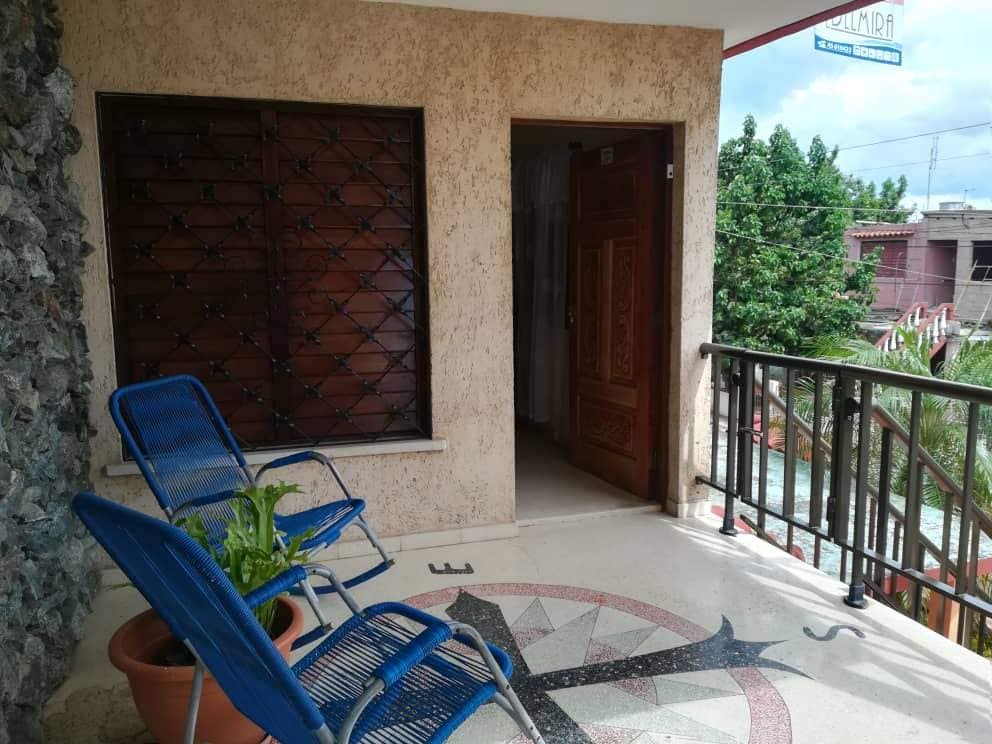 A private porch area is depicted with two blue chairs positioned next to a potted plant. A decorative tiled floor pattern is visible, while sturdy wooden railings frame the space, allowing for a view of greenery in the background.