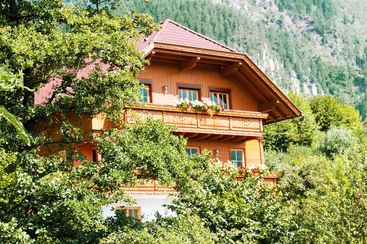 A three-story wooden house is surrounded by lush greenery, featuring a sloped roof and balcony with flower boxes. Large windows provide glimpses of the inviting interior, while the backdrop includes lush mountains, suggesting a serene natural environment.