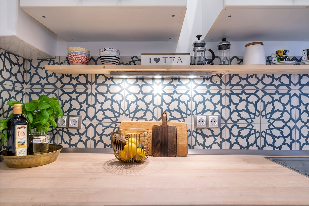 A modern kitchen space is highlighted with decorative tile backsplash and wooden shelving. On the countertop, a bowl holds fresh lemons, while a cutting board rests nearby. Various utensils and containers are neatly arranged, adding functionality to the inviting environment.
