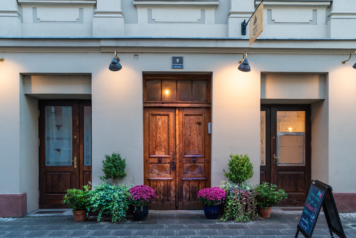 The image displays an inviting entrance adorned with rustic wooden doors, flanked by green shrubs and brightly colored flowers. Warm lighting highlights the architectural details of the door frames, creating a welcoming appeal to the entrance, which is situated along a charming street.