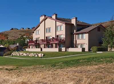 The image depicts a three-story building designed in a neutral color scheme, featuring multiple balconies on the upper floors. The surrounding green lawn contrasts with the natural landscape, which includes rolling hills in the background. Clear blue skies provide a bright backdrop.