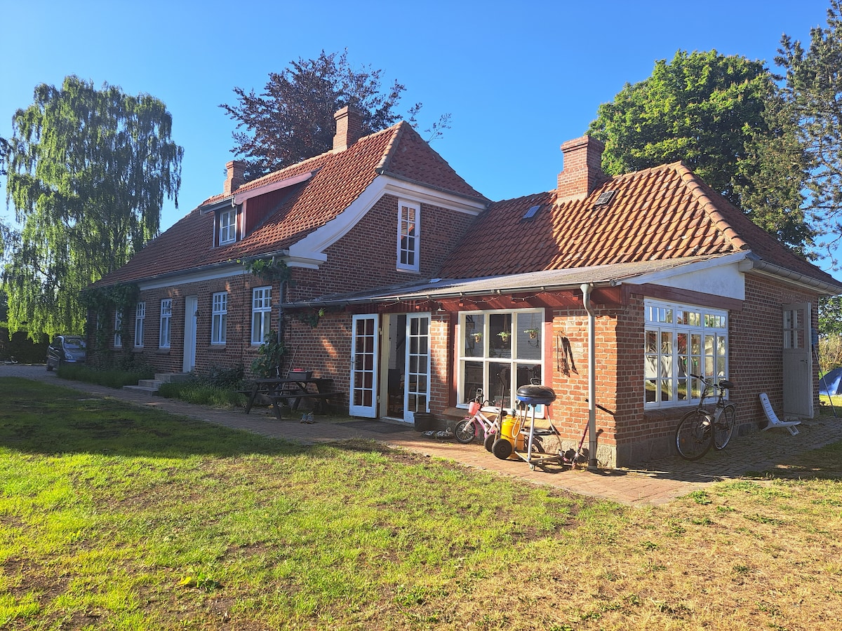 A charming brick house is showcased with distinctive rooflines and multiple windows allowing for abundant natural light. A spacious outdoor area features a patio with seating and bicycles nearby. Lush greenery surrounds the property, creating an inviting atmosphere for family gatherings.