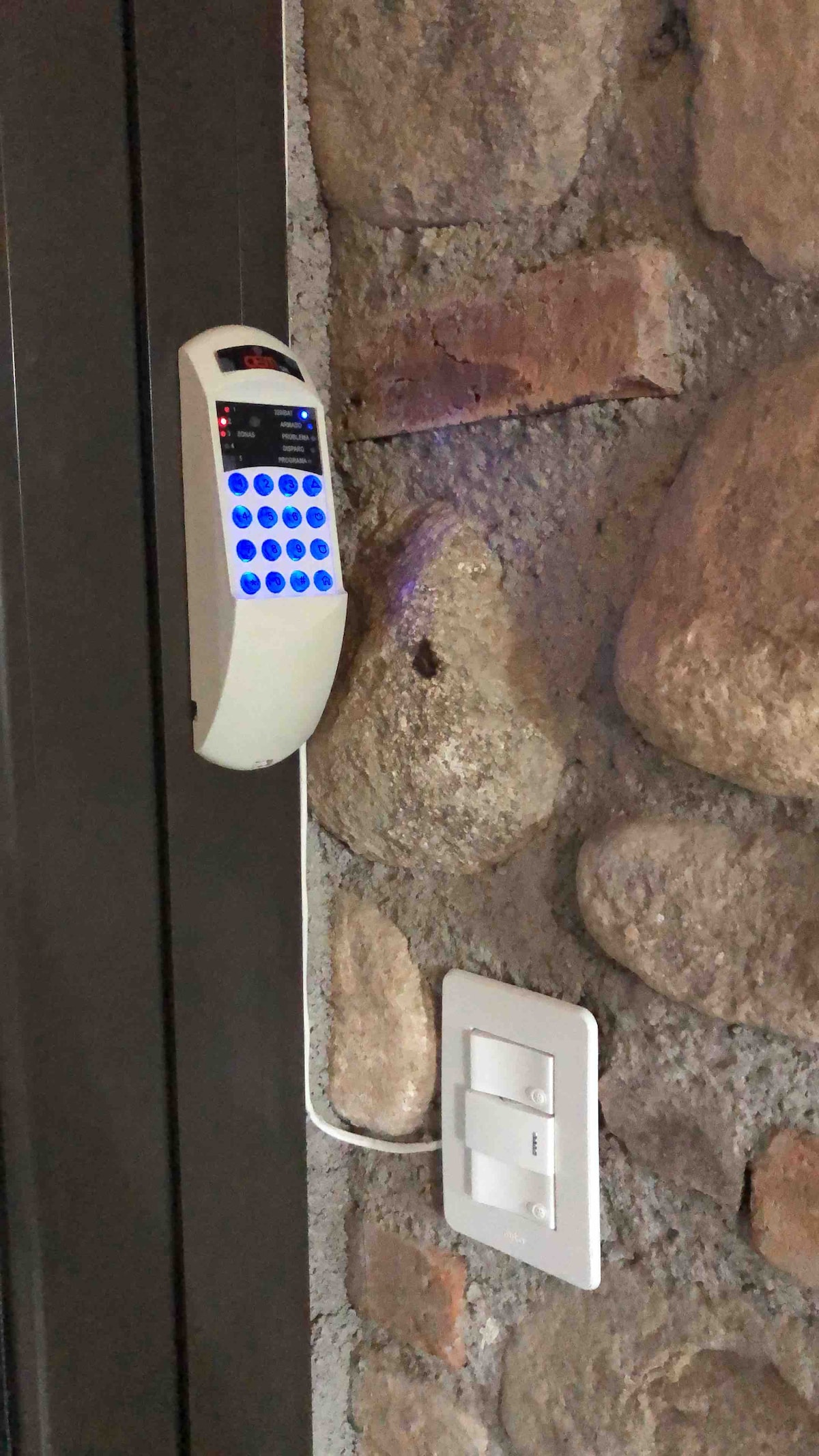 A security keypad is mounted on a rustic stone wall, featuring a digital display and a keypad with blue backlit buttons. A light switch is located below the keypad, providing a simple access point next to the textured natural stone.
