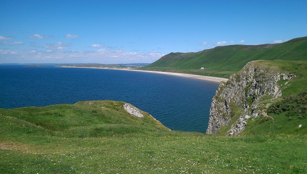 A panoramic view of a sandy beach curves along the coastline, bordered by lush green hills under a clear blue sky. The shimmering water meets the shore, while cliffs stand prominently along the landscape, creating a peaceful natural setting.