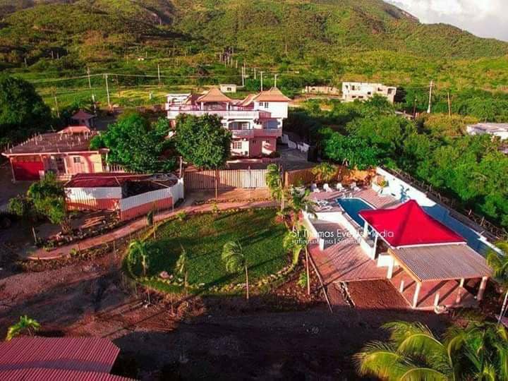 An aerial view captures the hotel complex surrounded by lush greenery, featuring a swimming pool and various outdoor patios. The building's architecture consists of multiple levels with red roofs and a mix of vibrant outdoor spaces, blending harmoniously with the natural landscape.