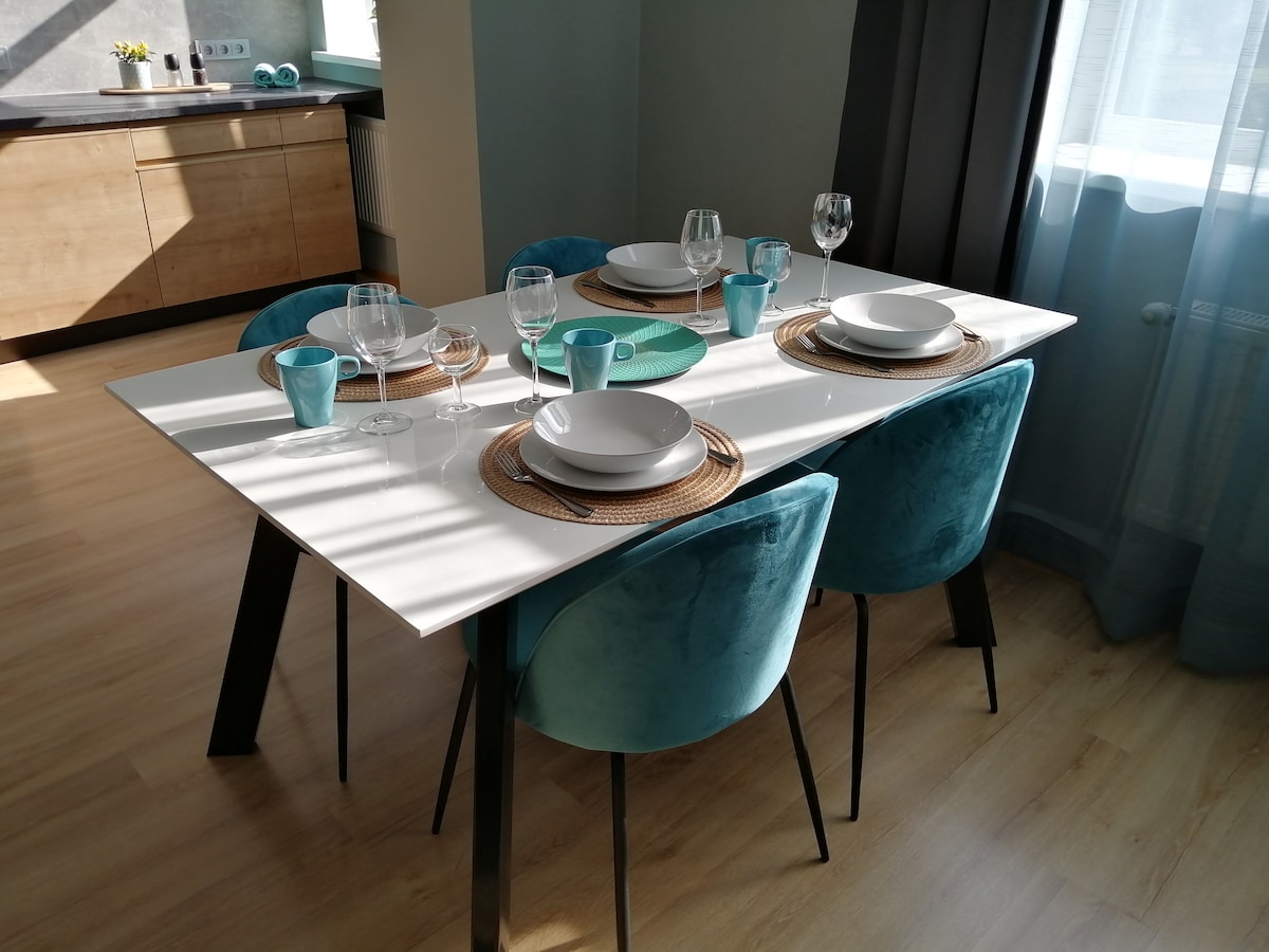 A dining table set for six features a white surface and dark legs, with turquoise and natural woven placemats. Glasses, plates, and cutlery are neatly arranged, complemented by sunlight streaming through a nearby window, casting soft shadows on the wooden floor.