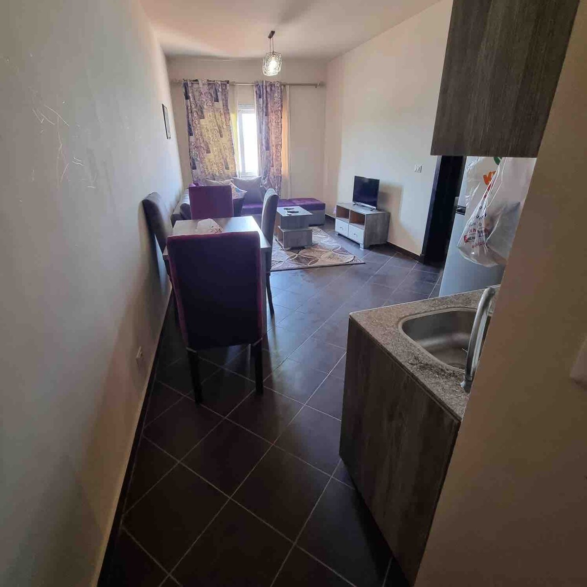 A cozy living area is highlighted, featuring a seating arrangement with two purple chairs and a small table. Natural light is invited through a window adorned with patterned curtains. A television and a kitchenette are visible, complemented by dark tiled flooring throughout.