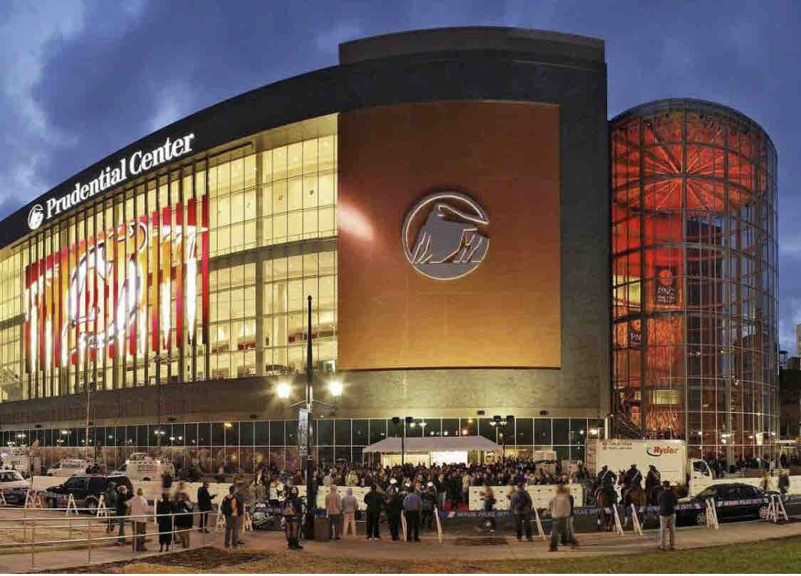 The Prudential Center is displayed in an evening setting, showcasing its large glass façade illuminated with colorful lighting. A crowd gathers in front, with the arena’s logo prominently visible. The structure’s modern design features a cylindrical entrance, contributing to its architectural appeal.