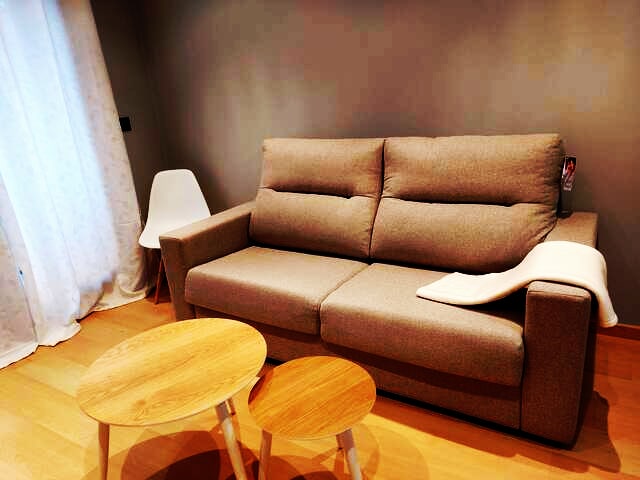 A comfortable sofa with plush cushions is positioned in a softly lit living area. Two small, round wooden tables are arranged in front of the sofa. A floor-to-ceiling window with sheer curtains allows natural light to filter in, enhancing the overall warmth of the space.